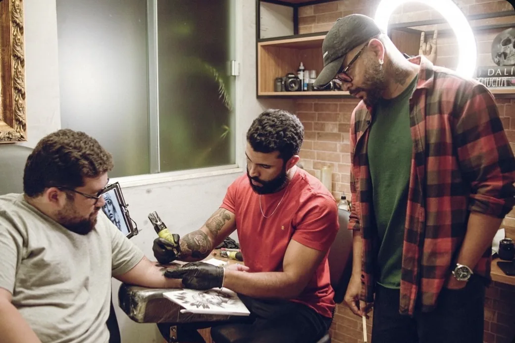 Matheus tatuando o antebraço de um cliente no estúdio, com a arte impressa ao lado