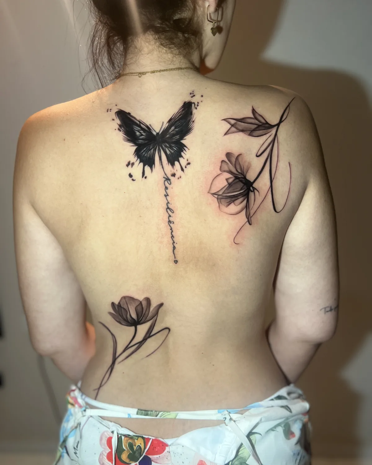 Tatuagem feminina nas costas com borboleta preta, flores em raio-x e a palavra Resiliência em caligrafia