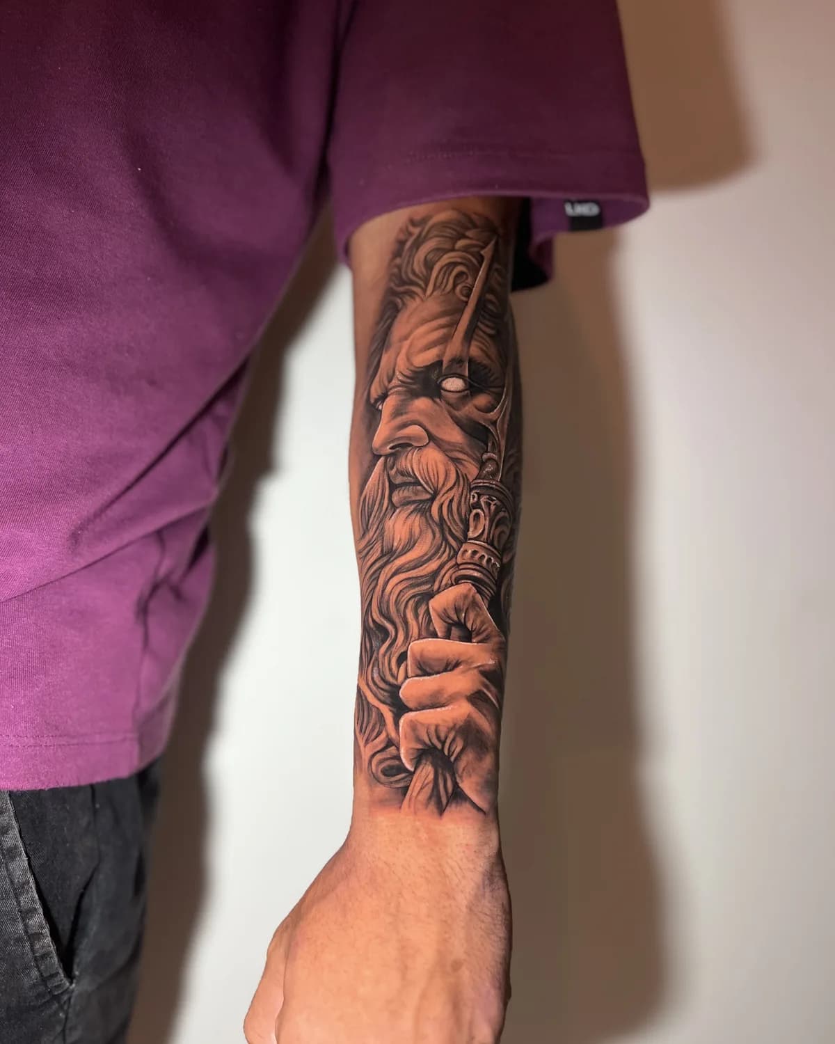 Tatuagem realista de Zeus segurando um raio, em preto e cinza no antebraço